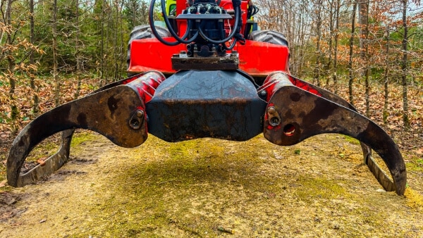 A machine with a claw attachment is placed on a dirt path surrounded by trees and fallen leaves in daylight.